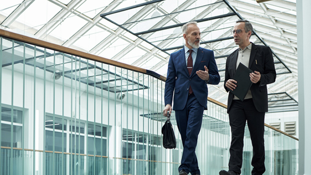 Two business professionals in suits walking through a glass covered office walkway while talking and carrying work bags and documents.
