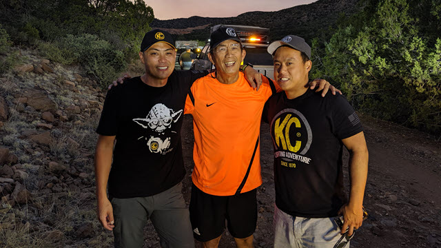 Three men, Mike Wang (center) and his sons Alan (left) and Andy (right), stand together on a scenic overlook at dusk.  