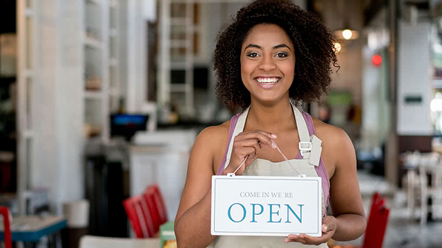A smilling woman holding a sign that reads come in we're open.