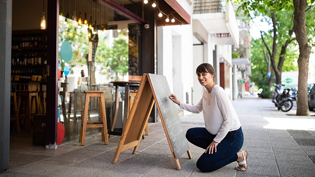 A woman kneeling and writing on a sandwich board.